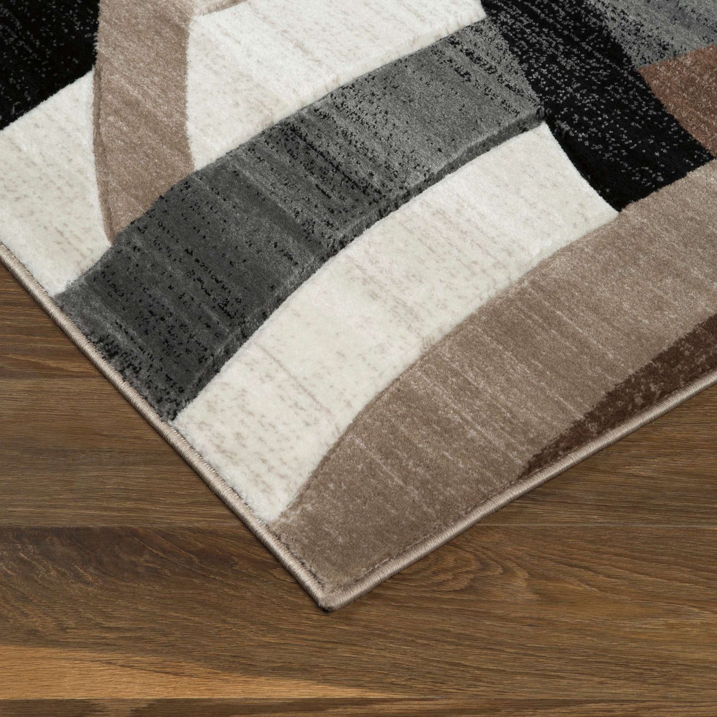 A close-up of a modern area rug with an abstract geometric pattern in beige, brown, gray, black, and off-white, shown on a dark wood floor.