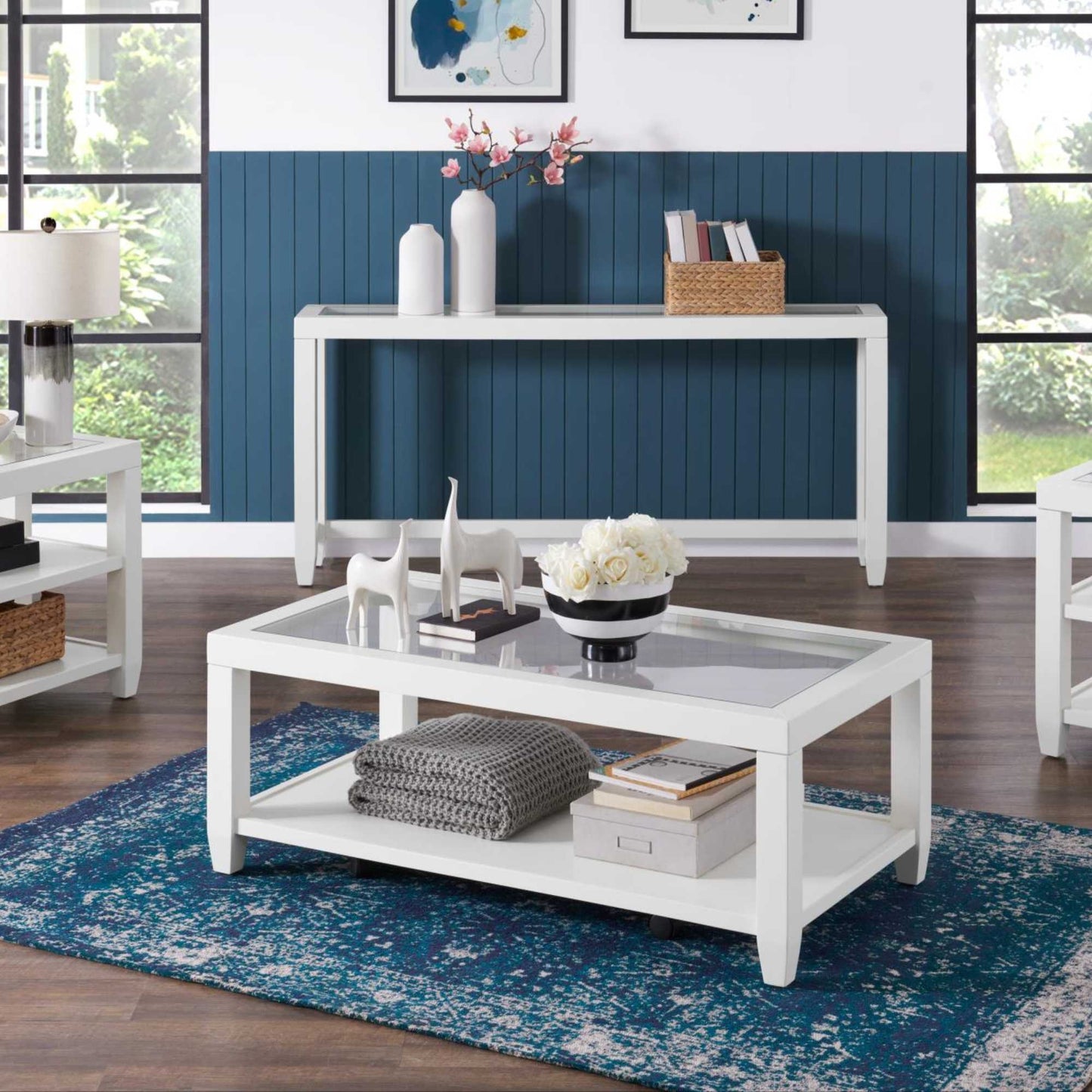 A white rectangular coffee table with a glass insert top and a display bottom shelf, placed on a blue area rug in a room with a blue wall and a window.