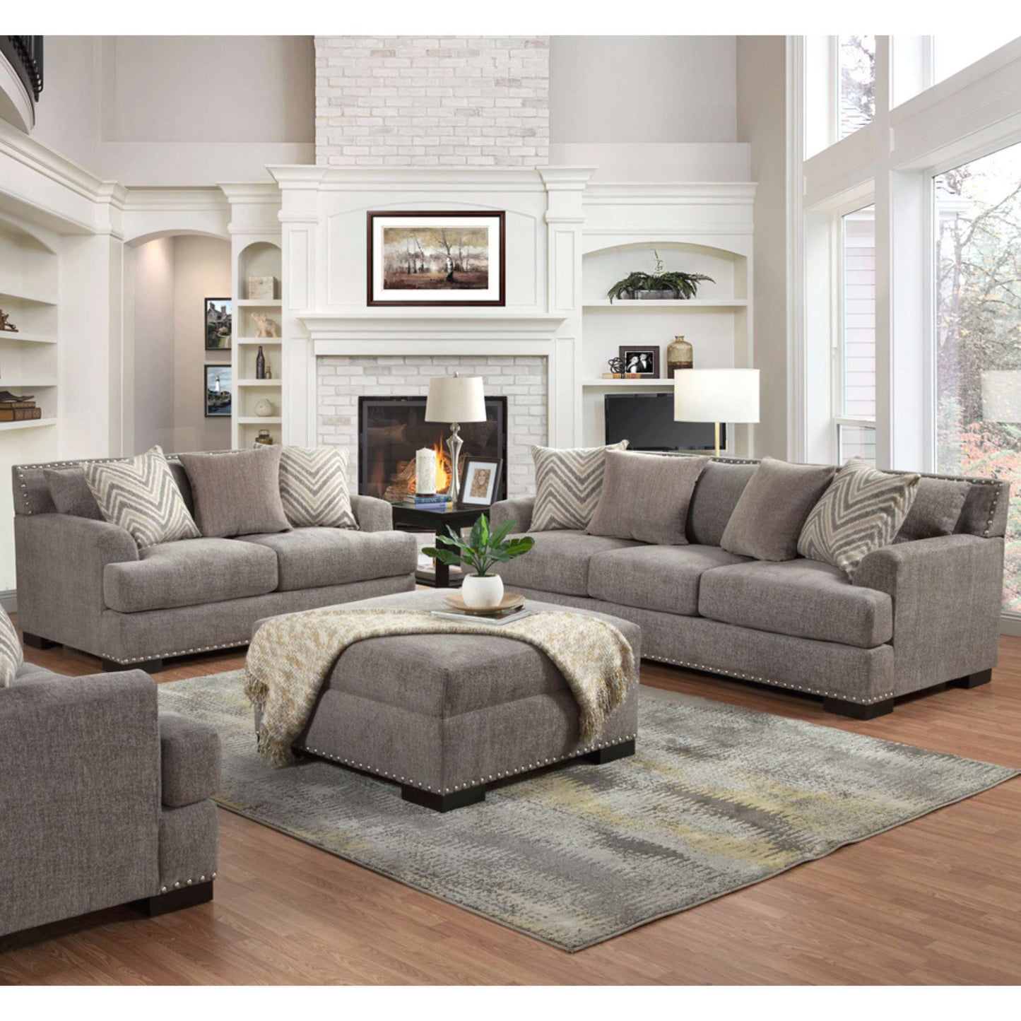 A set of granite grey upholstered sofas and loveseat with decorative nailhead trim, arranged in a living room setting with a fireplace, bay window, and decorative pillows.
