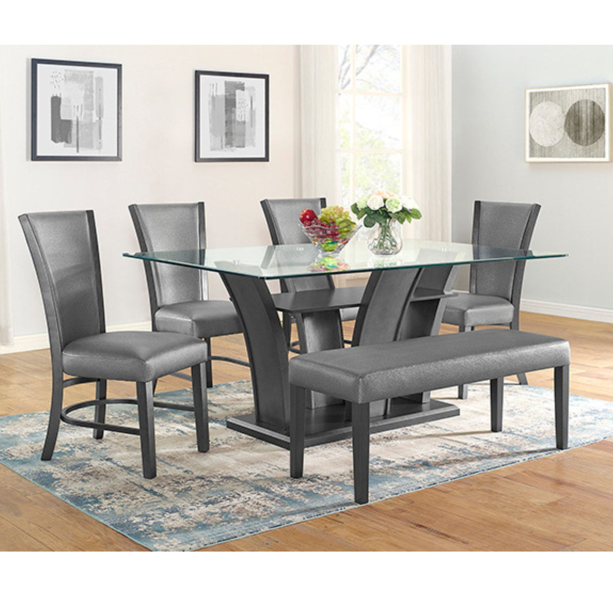 Winsor 6-piece gray dining room set featuring a rectangular glass-top table with a sculptural pewter base, four high-back gray upholstered chairs, and a matching gray bench, staged on a blue patterned rug in a bright dining room.