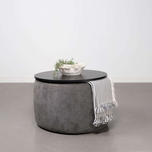 Round gray ottoman with a black top and a white bowl with greenery on a light gray floor.