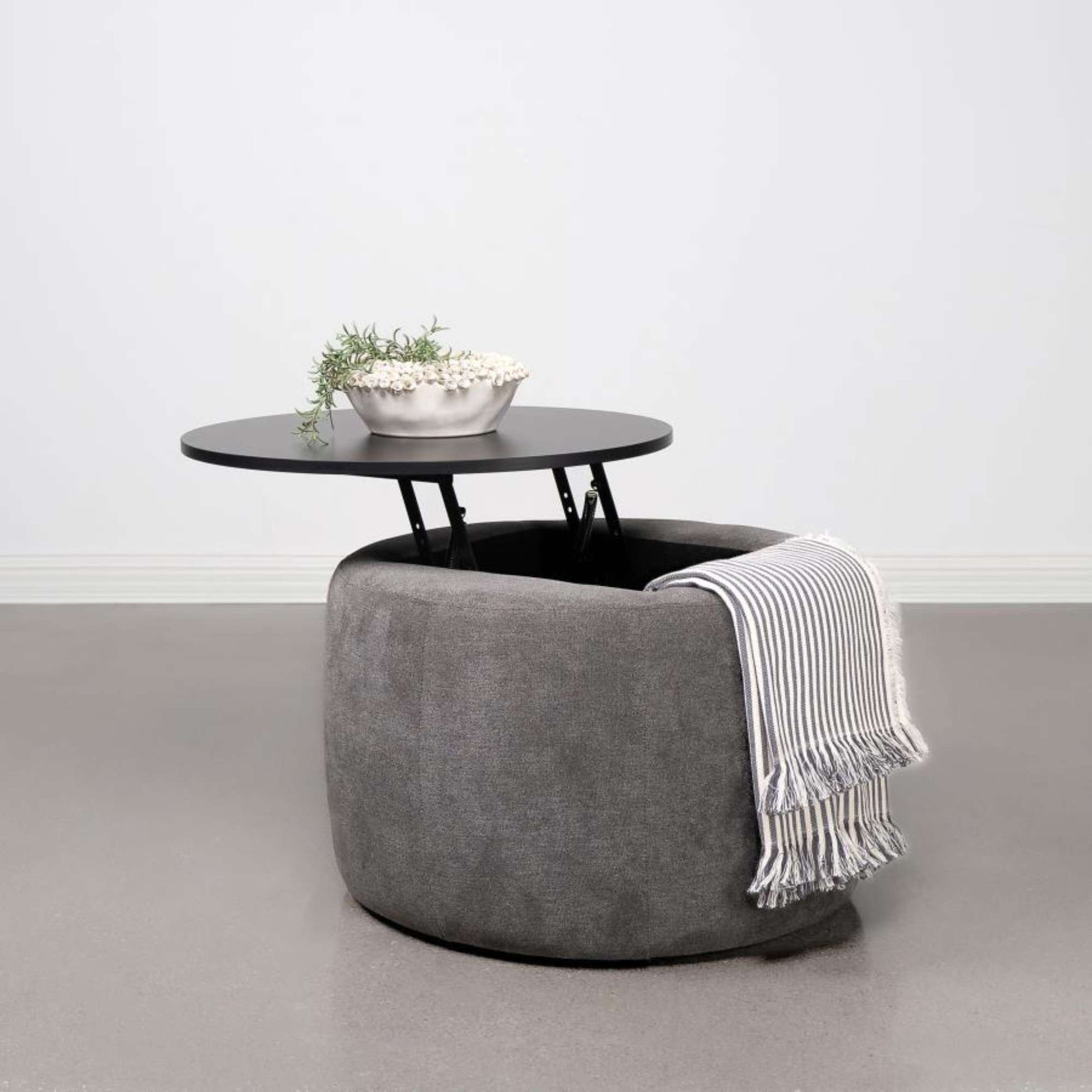 Gray ottoman with a black lift-top table and a white and gray striped blanket on a light gray floor.