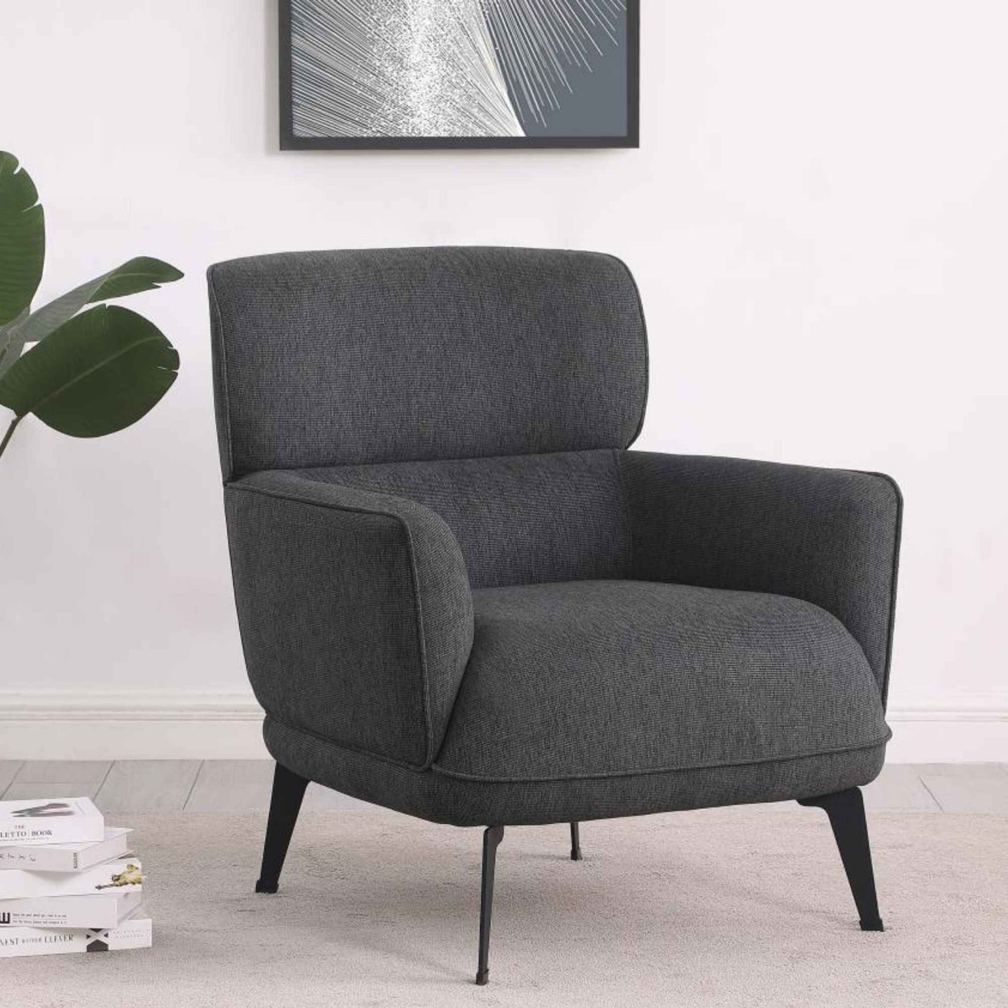 Gray armchair in a room with a plant and books on a white wall.