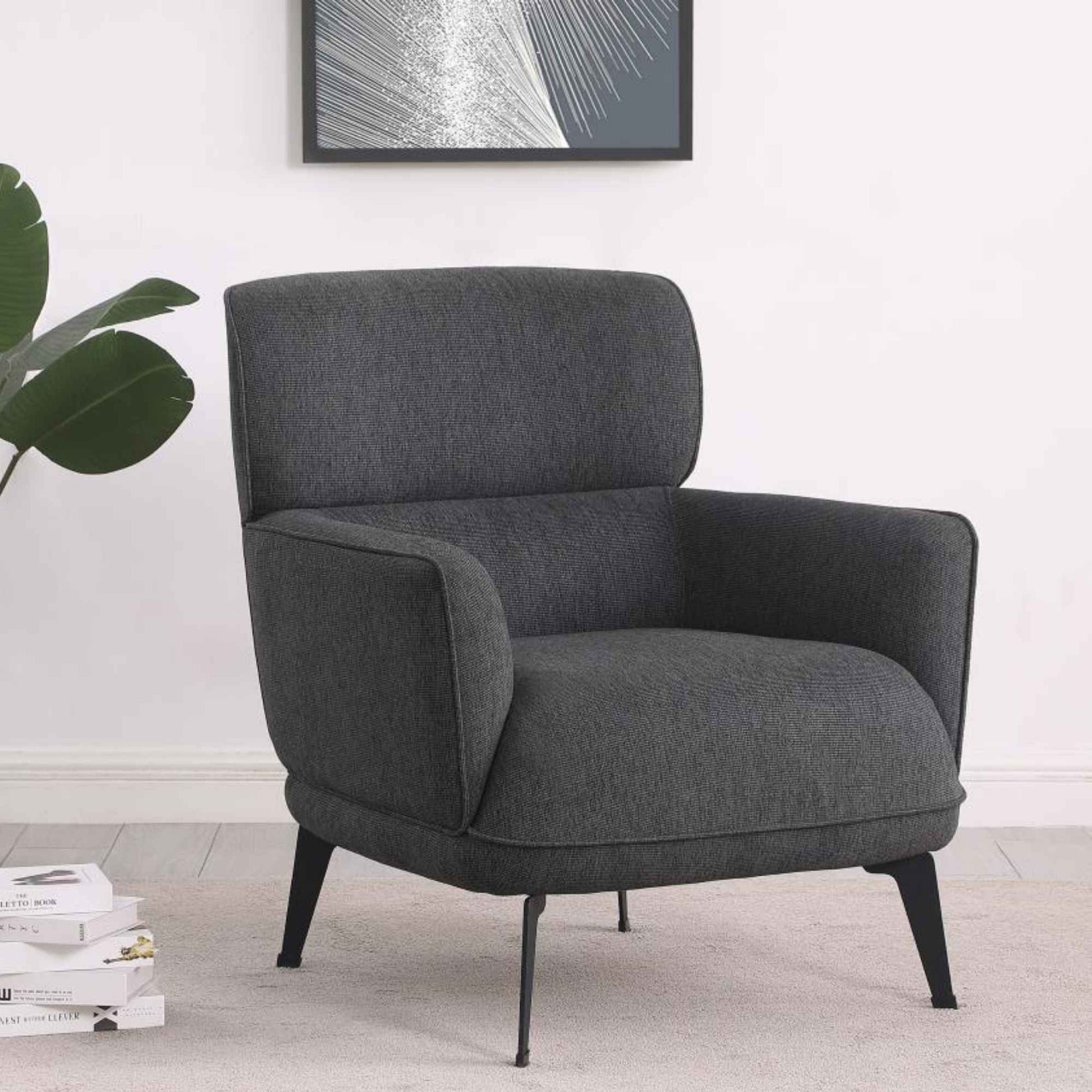 Gray armchair in a room with a plant and books on a white wall.