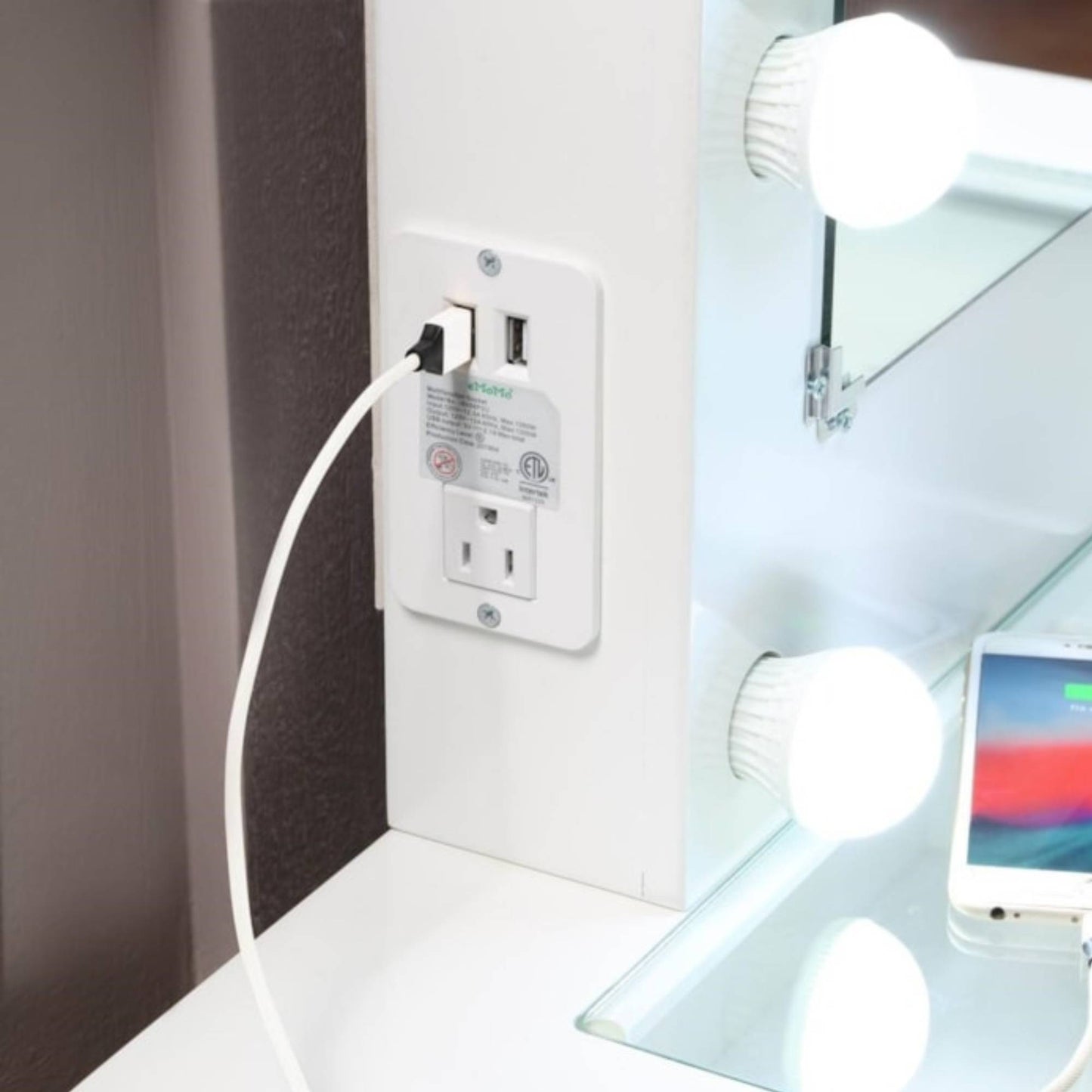 White LED light bulb being plugged into a wall socket with a phone on vanity table in the background.