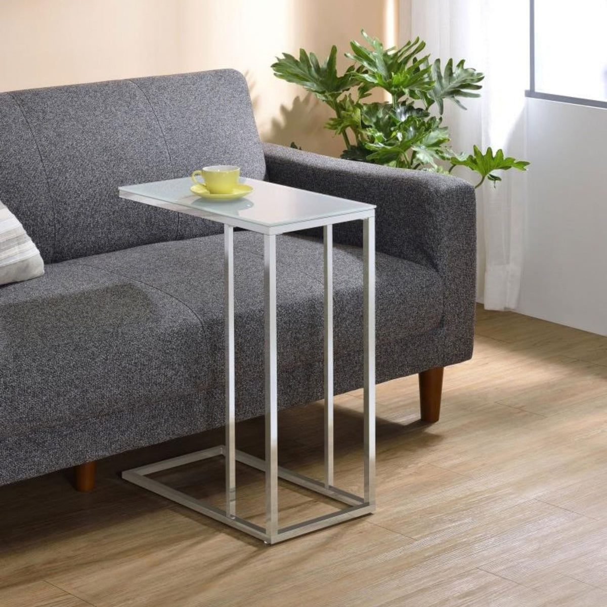 Jurgen modern silver metal side table with a glass top, shown in a living room setting next to a gray fabric sofa with a yellow cup on the tabletop.