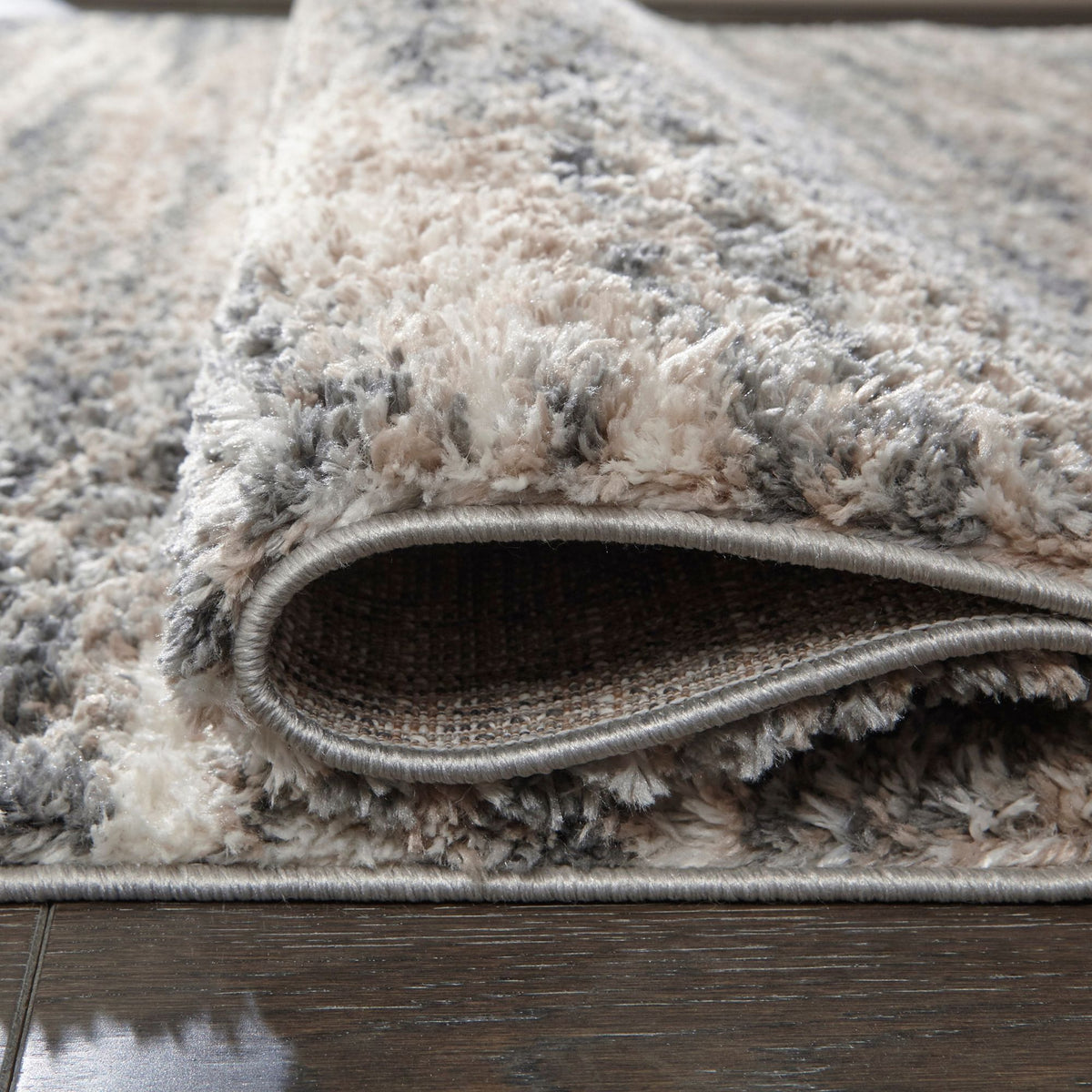 Close-up of a plush R404861 area rug with a mixed neutral pattern in beige, gray, and cream, folded over to show the underside and edge, placed on a dark wood floor.