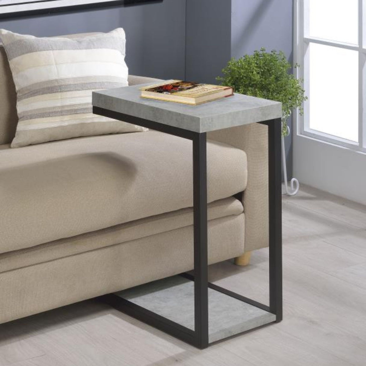 Wendral C-Shape Accent Table in Faux Cement veneer finish with black finished legs in living room setting with book on top. 