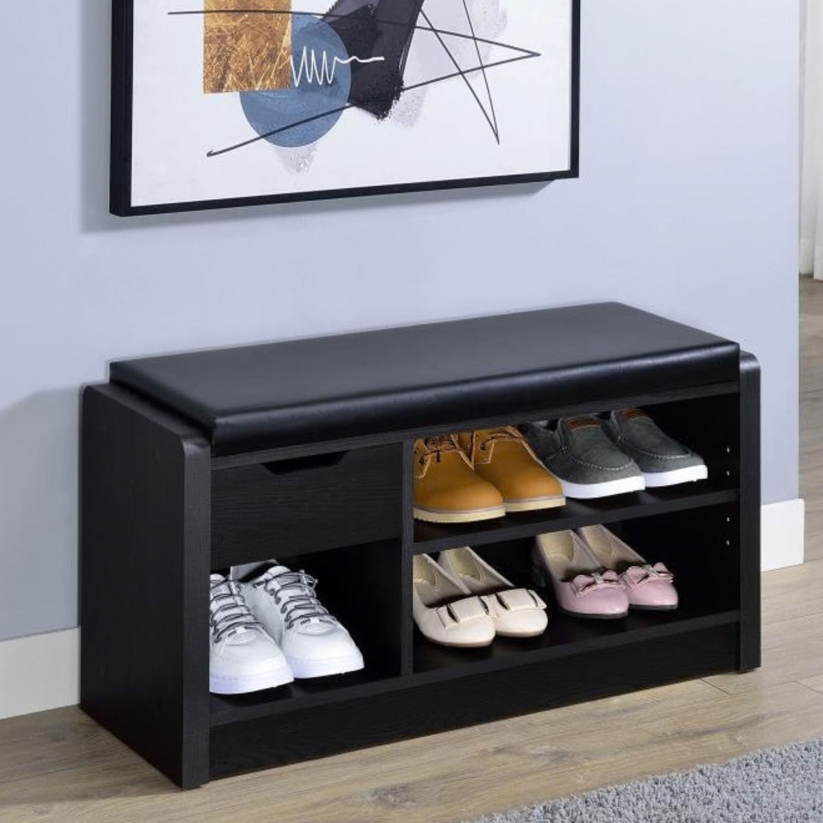 Yumia black wooden shoe bench with a padded top, shown in a room setting with various shoes stored in its open compartments. A framed abstract print hangs on the light gray wall above the bench.
